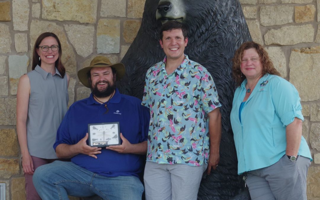 Four people in front of a bear statue. The second person from the left is holding an award.